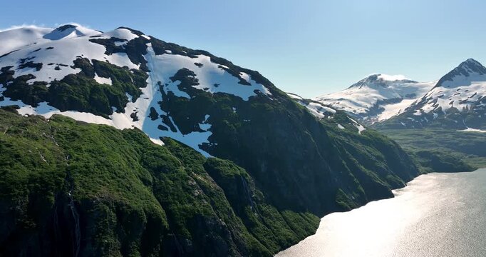 Verdant mountains covered with show on the tops. Bright sun dazzles the rocks and the surface of the lake. Alaska, USA.