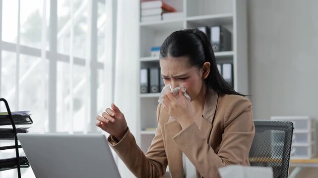 Professional asian businesswoman working on laptop while battling seasonal flu, experiencing nasal congestion and discomfort in clean, modern office environment
