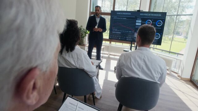 Group of diverse doctors wearing white coats listening to young male lecturer during professional development course in modern hospital