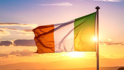 Irish flag waving on flagpole with gold finial, vibrant sunset sky in background, symbol of national pride and unity.