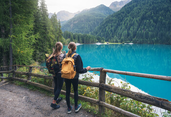 Two young women with backpacks standing near wooden fence overlooking turquoise alpine lake in the Dolomites mountains, Italy. Scenic reflection view with girls, trees and lake. Hiking and trekking