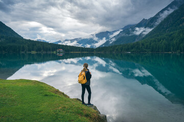 Woman with yellow backpack standing near lake, looking at reflection of mountains and forest in calm water in fall. Cloudy sky and autumn colors create a scenic alpine view. Hiking in Dolomites, Italy