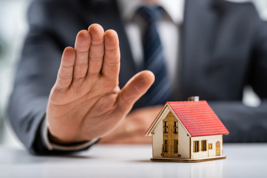Businessman in a suit making a stop gesture with hand behind a small detailed house model symbolizing real estate protection and property rights concepts
