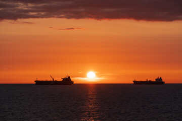 Two silhouette industrial oil tankers sailing on the tranquil Baltic Sea against a vibrant orange and red sunset sky.