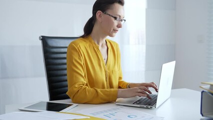 Professional businesswoman wearing yellow shirt and eyeglasses working intently, typing on laptop in bright office space with documents and tablet surrounding workspace. Business people concept - Powered by Adobe