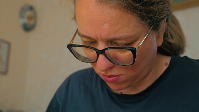 Close-up of a woman's face with glasses, focused on something of her own, in a casual black T-shirt. The background is made in soft textures and muted colors. A woman is busy embroidering