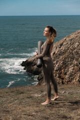 Female yoga practitioner in a sleek bodysuit stands on a rocky coastline, holding a yoga mat, gazing at the ocean, embodying tranquility and connection with nature