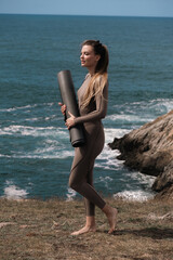 Athletic woman in form-fitting activewear stands on rocky shore, holding yoga mat, with ocean waves crashing in the background, embodying fitness and tranquility