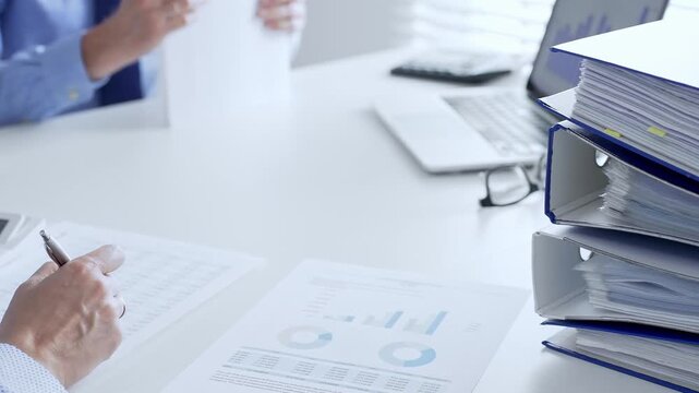 Corporate professionals reviewing financial documents, working collaboratively at white desk with blue folder stack, highlighting teamwork and business productivity. Audit and taxes in business