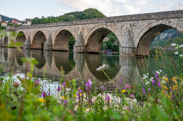 Fototapeta premium An ancient stone bridge with arches spanning the tranquil Drina River with wild flowers in the foreground