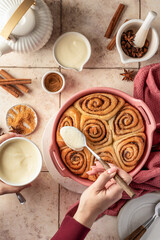 Female hands pouring glaze over freshly baked cinnabon buns in ceramic baking dish, beige tile background. Making cinnamon rolls process, top view