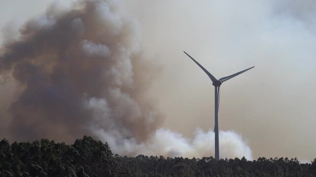 A dramatic wildfire burns near a windmill under a sky filled with thick clouds. The contrast between renewable energy and destructive flames highlights themes of climate change, environmental crisis