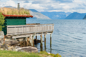 A green, wooden dock and boathouse with peat, thatched roof with chimney, and full wood deck, on wooden pillar stilts in the water of the coastline of the Sognefjord in Norway, Europe