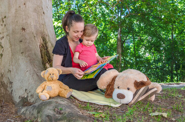 Mother reading a book with her cute little child under an old plane tree in the park. Funny and educative outdoor activity for children, in sunny summer day