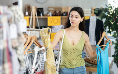 Interested Armenian woman in a knitted top stands in a clothing store and chooses summer dresses on...