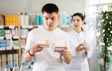 In sales area of pharmacy, guy examine packaging of tablets pills, reads characteristics and properties, checks expiration date, storage conditions. Girl pharmacist offers analogue and additional drug