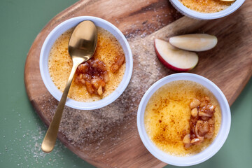 Creamy dessert featuring caramelized sugar topping, served in white ramekins on a wooden board, garnished with fresh apple slices and brown sugar, showcasing culinary artistry