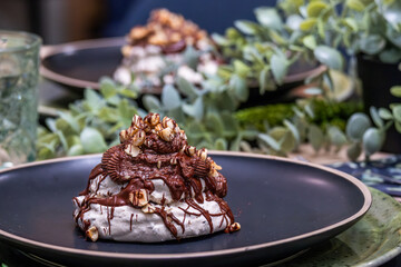 Dessert featuring a meringue topped with chocolate drizzle, popcorn, and cookie pieces, elegantly presented on a black plate surrounded by greenery and soft lighting