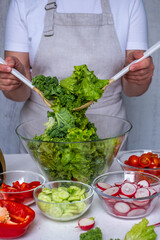 Hands of a person mixing fresh green salad in a large glass bowl surrounded by colorful vegetables, showcasing healthy eating and vibrant culinary creativity