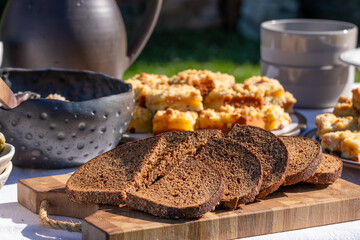 Freshly sliced dark bread on wooden cutting board, surrounded by delicious baked goods and rustic pottery, creating a warm and inviting outdoor dining atmosphere