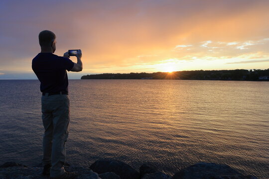 Middle-aged man taking a picture from his cell phone of the sun rising over the water - Powered by Adobe