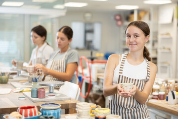 Teen girl visitor to course on creating ceramic products shows off final product, shows painted bowl. She holds small ceramic vase in her hands, smiles and rejoices at successful training