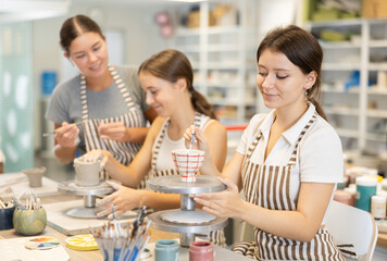 Young female student painting ceramic cup with brush in workshop