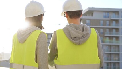 Two architects wearing safety hard hats and vests holding blueprint and discussing construction site project at sunrise, back view. Architecture, engineering and teamwork concept