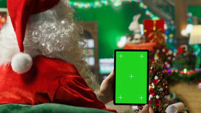 Man dressed as Santa Claus bringing joy to others during holiday season, posting online on green screen tablet. Santa Claus using chroma key device to spread sense of wonder during Christmas, camera A
