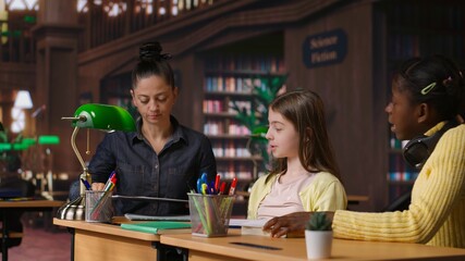 Caucasian tutor guiding elementary school girls at a library desk, providing private lessons to aid in academic development with work books and notes. Teacher discussing with scholars. Camera B.