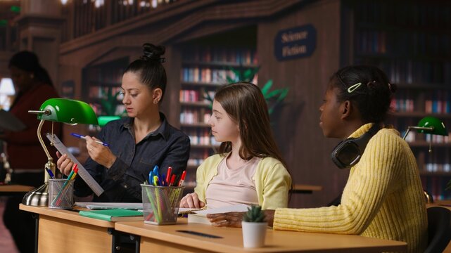 Elementary school pupils working with a professor solving exercises for the class homework, developing learning skills at a study desk. Tutor explains the curriculum materials to girls. Camera B.