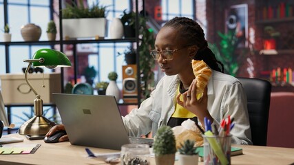 Busy freelancer in home office eating snack on the go while typing on laptop keyboard. Woman enjoying croissant while using electronic device to solve job tasks online, camera B