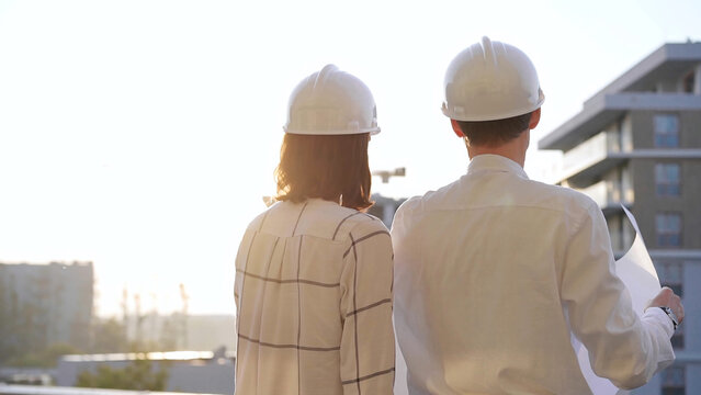 Man and woman architects constructor engineers wearing safety glasses and hardhats, are examining blueprints at a construction site during a sunset, back view. Architecture concept