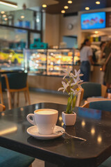 Cup of cappuccino sits on a dark table in a cozy cafe with a small white flower decoration. Concept of coffee break, cozy atmosphere, urban lifestyle