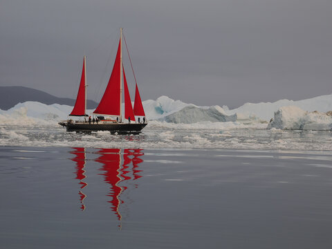 Red-sailed boat sailing through icy arctic waters surrounded by floating ice and large icebergs under cloudy sky.