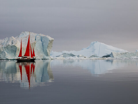 Red-sailed boat sailing through icy arctic waters surrounded by floating ice and large icebergs under cloudy sky.