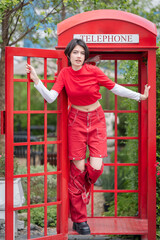 A pretty young woman in a modern red outfit is inside a red public phone booth, holding a vintage pay phone. A cute teenage girl expresses a variety of emotions while talking.