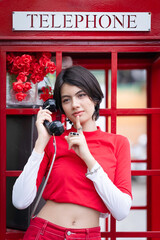 A pretty young woman in a modern red outfit is inside a red public phone booth, holding a vintage pay phone. A cute teenage girl expresses a variety of emotions while talking.