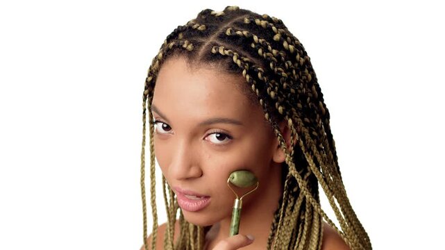 A young African American woman is using a jade roller on her face, smiling while pampering herself in a bright, well-lit environment, showcasing her commitment to beauty and skin care.
