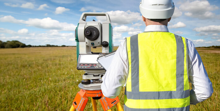 Surveyor in hard hat and safety vest using a total station for land measurement in an open field. Activity: Land Surveying, Construction, Geodesy.