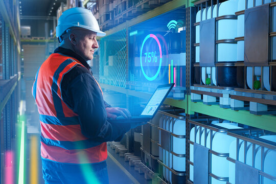 Worker in helmet and safety vest using laptop for smart warehouse inventory management with digital data analytics. Logistics, Technology, Warehouse.