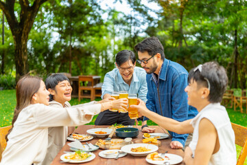 Group of happy diverse Asian millennial people friends enjoy holiday celebration social gathering dinner party in garden evening, drinking beer and toasting glasses together outdoor festive event.