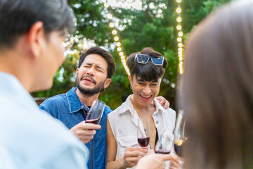Happy diverse Asian millennial people friends enjoy meeting holiday celebration social gathering dinner party in garden evening, eating food, drinking wine and talking together outdoor festive event.