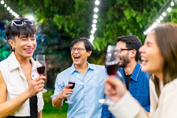 Happy diverse Asian millennial people friends enjoy meeting holiday celebration social gathering dinner party in garden evening, eating food, drinking wine and talking together outdoor festive event.