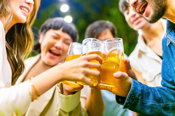 Group of happy diverse Asian millennial people friends enjoy holiday celebration social gathering dinner party in garden evening, drinking beer and toasting glasses together outdoor festive event.