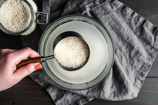 Hand Holding a Measuring Cup of Soaking Sticky Rice: Scoop of glutinous rice held over a glass bowl full of cloudy water and soaked sweet rice