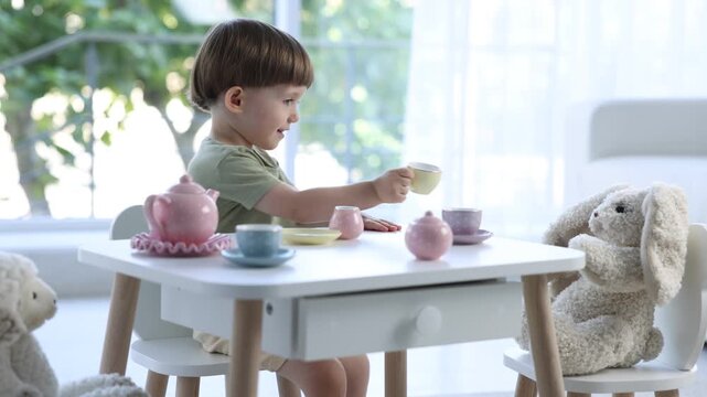 Cute little boy playing tea party with his toys at table indoors