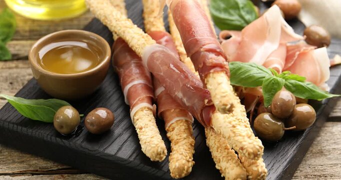 Woman taking grissini breadstick with prosciutto from board at wooden table, closeup
