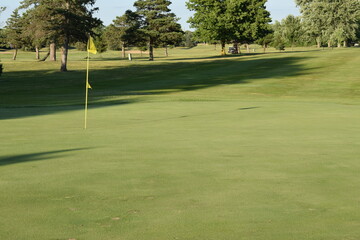 Golf course putting green and partial fairway view showing the goal of sinking a put in the location indicated with the yellow flag and pole.
