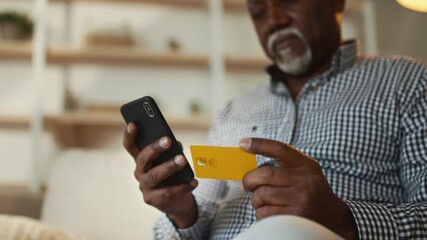 An elderly African American man sits comfortably at home, focused on his smartphone while holding a credit card. He appears engaged and relaxed in his personal space. - Powered by Adobe